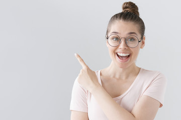 Horizontal picture of young good-looking female isolated on grey background wearing big round spectacles, pointing leftwards with forefinger, looking extremely happy, satisfied and surprised.