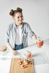 Fototapeta na wymiar Happy girl with buns smiling stretching glass with grapefruit detox smoothie to somebody. White wall background. Healthy diet food.