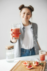 Happy girl with buns smiling stretching grapefruit detox smoothie to  camera over white wall. Healthy diet food. Focus on glass.