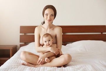Happy naked young mother smiling hugging her newborn nice baby sitting on bed together.