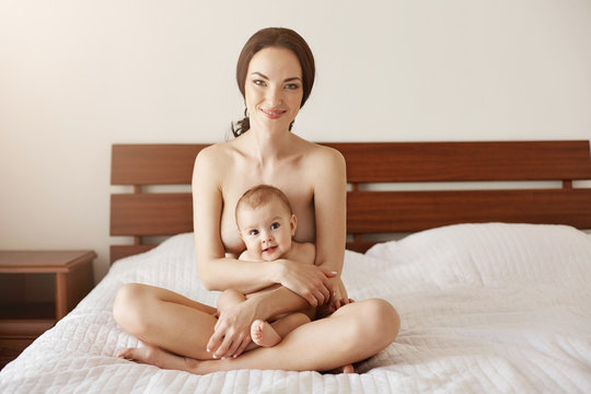 Happy naked young mother smiling hugging her newborn nice baby sitting on bed together.