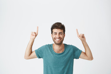 Fototapeta premium Check this out Cropped studio shot of attractive good-looking excited young man in blue t-shirt pointing fingers up having surprised look, with cheerful and happy expression of face. Broadley smiling.