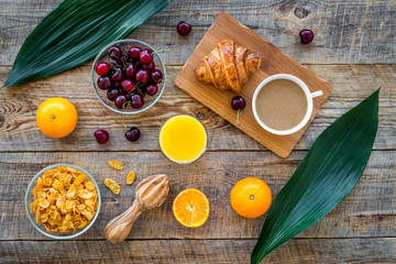 Light tasty breakfast. Muesli, oranges, cherry, french croissant and milky coffee on wooden table background top view