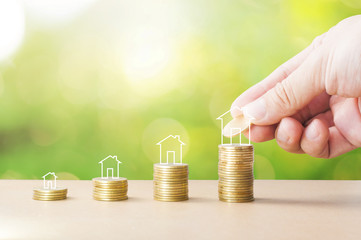 Hand adding coin to stack of golden coins with house icon on wooden table with bokeh green...