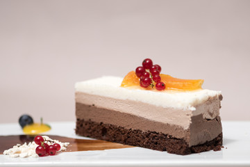 Fancy triple mousse chocolate dessert, placed on a white plate, decorated with forest fruits and chocolate sauce, white plate, light brown background