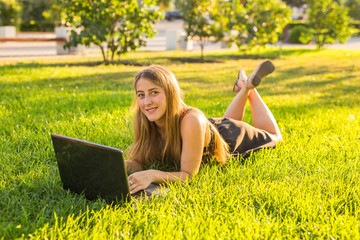 portrait of a beautiful student girl with laptop computer on the green grass in the park in spring...