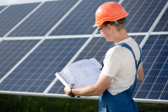 Young worker in overall looking in drawings at solar power station. Employee in blue overall and orange helmet, looking in technical documentation.