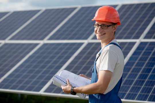 Young man working with solar panels. Worker at solar power station holding documents looking in camera and smiling.