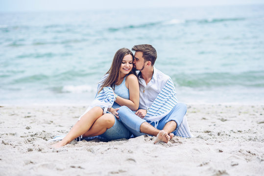 Beautiful pregnant woman and her respectable man leaning against each other and covered with a white striped plaid sitting on the seashore. Lovely young couple.