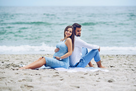 Beautiful pregnant woman and her respectable man leaning against each other and sitting on the seashore. Lovely young couple.