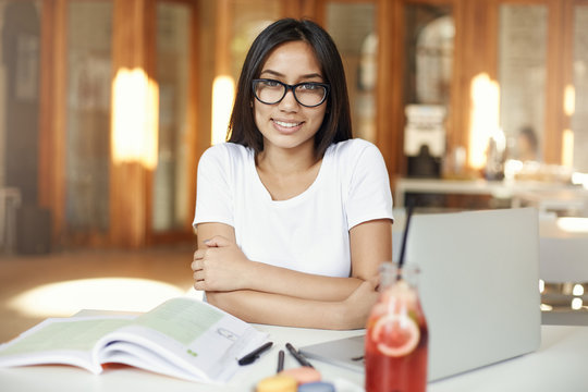 Portrait of assian woman smiling working on laptop in cafe or bright coworking space wearing glasses.
