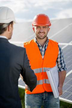 Foreman shaking hand to a business client. Worker in orange helmet and vest with a client in business suit an white helmet, solar panels at backstage.