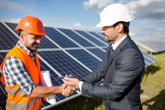 Businessman holding photovoltaic detail and shaking hand to a foreman. Solar panels in the field, business deal between client and foreman.