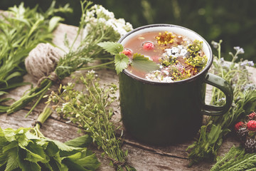 Summer healthy herbal tea in old enameled mug and bunches of healing herbs on wooden board. Herbal...