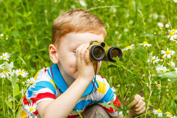 Red-haired boy is playing: sitting in an ambush looking at toy binoculars on a background of a...