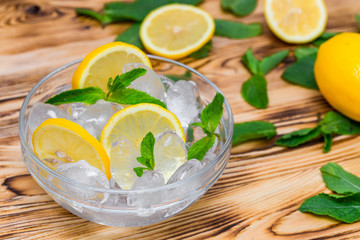 Fresh sliced lemon, bright green mint leaves and frozen ice cubes in a transparent bowl on a wooden table