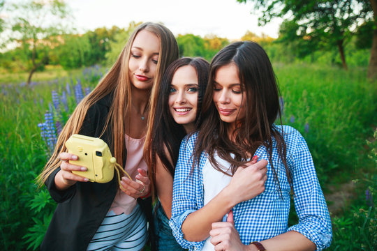 Three hipsters girls blonde and brunette taking self portrait on polaroid camera and smiling outdoor. Girls having fun together in park.