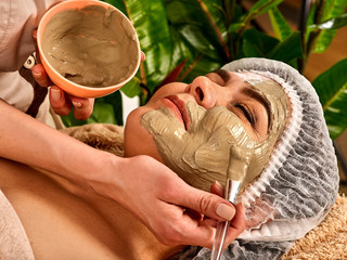 Mud facial mask of woman in spa salon. Massage with clay face in therapy room. Applying beautician with bowl therapeutic procedure on green plants background. Therapeutic mud used in spa procedures.