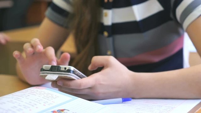 Schoolgirl dressed in a jacket sits at a school desk and holding a silver smartphone
