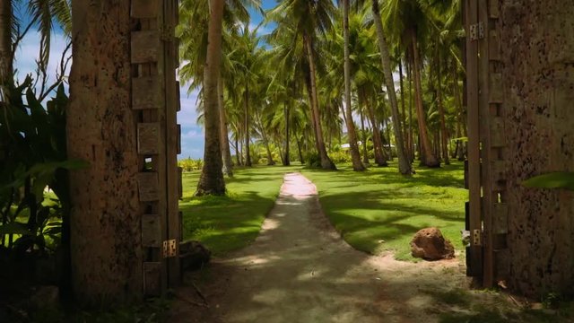 Camera moving through rustic gates to park with palm trees on lawn grass, Philippines