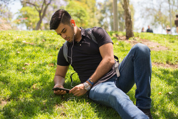 Naklejka na ściany i meble Male college student lying in grass on campus, looking at his cell phone; wearing backpack and earphones