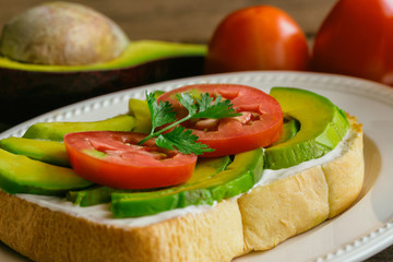 Fototapeta na wymiar Open sandwich style for breakfast or lunch. Sandwich spread with cream cheese, avocado, tomato and parsley on white plate. Avocado open sandwich style serve with orange juice on rustic wood table.