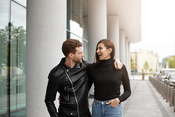 Stylish confident young bearded male wearing black leather jacket hugging beautiful laughing lady, trying to impress her, telling funny stories and jokes during nice walk outdoors at weekend