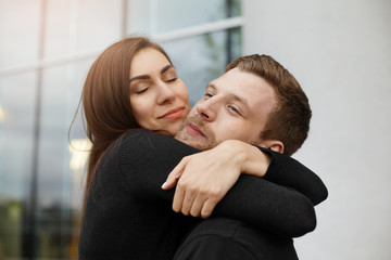 Happy beautiful young Caucasian couple cuddling outdoors. Beautiful brunette woman closing eyes in pleasure while hugging her bearded boyfriend after long separation. Love and happiness concept