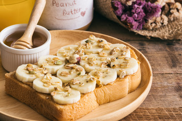 Banana peanut butter toast sandwich on wood plate with honey. Homemade open sandwich spread with peanut butter on top with banana slice and granola. Delicious healthy sandwich for breakfast or lunch.