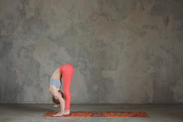 Young woman practicing Standing forward bend, Uttanasana yoga pose against texturized wall / urban...