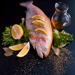 Fresh trout and ingredients to prepare fish dishes on black table, with spices and lemon wedges, top view