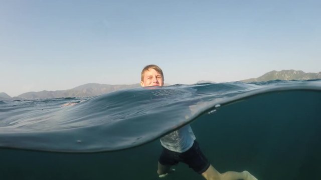 young man in a white shirt is swimming on the sea, half underwater close up, raw, 4k