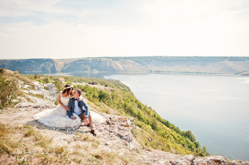 Stunning young wedding couple sitting on the edge of the cliff with a beautiful scenery on the background.