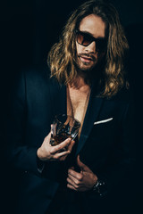 Handsome bearded long haired man in sunglasses and stylish suit holding glass of whiskey on black
