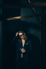 Stylish long haired man in suit and sunglasses holding glass while posing on black