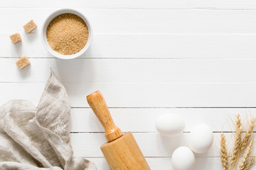Baking ingredients: brown sugar, white eggs, rolling pin, and wheat on white wooden table. Top...
