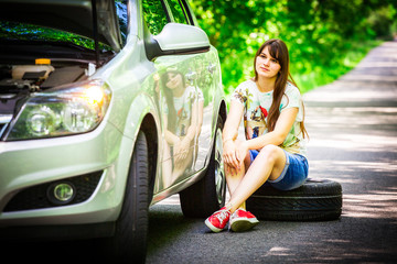 Young brunette woman sitting near a silver car on the roadside with a broken wheel