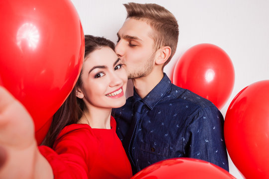 handsome guy kisses his pretty girlfriend on the cheek and making selfie on a white background
