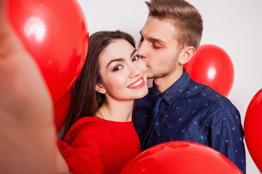 handsome guy kisses his pretty girlfriend on the cheek and making selfie on a white background