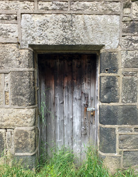 old faded plank bare wooden door in a stone building with grass and weeds growing over the bottom