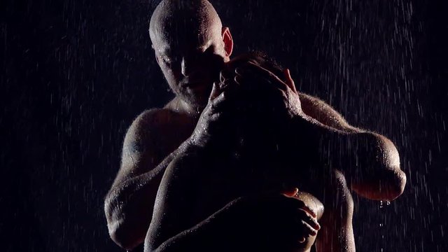 large muscular bald man in a dark room under the jets of water manipulates the head with his women. they are naked and wet. the pre-sex caresses.