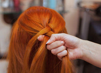 Fototapeta premium Beautiful girl with red long hair, hairdresser weaves a French braid, in a beauty salon. Professional hair care and creating hairstyles.