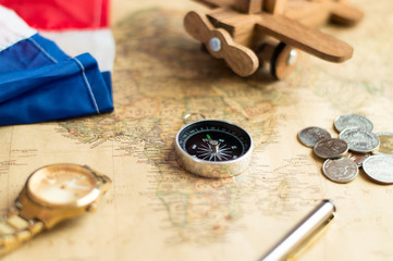 Fototapeta na wymiar Compass on vintage world map with coins, pen, wrist watch, plane and flag for vacation and travel concept, selective focus.