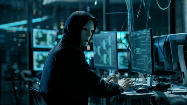 Masked Hacktivist Organizes Massive Data Breach Attack on Corporate Servers. He is in Underground Secret Location Surrounded by Displays and Cables.