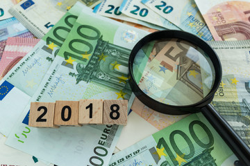 financial tax concept as magnifying glass on pile of euro banknotes and number 2018 on wooden block