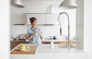 Beautiful ginger Caucasian woman standing at kitchen and enjoying morning coffee.