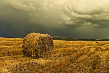 A round haystack on a cleared crooked field and a stormy sky. Beautiful landscape, space, hills in the background. After harvesting.
