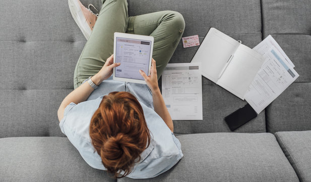 From above photo of ginger Caucasian woman sitting on couch and dealing with bills using tablet.