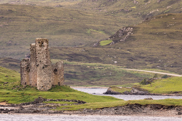 Assynt Peninsula, Scotland - June 7, 2012: Brownish Ruins of Castle Ardvreck from across Loch Assynt. Green ground vegetation. Brown mountain slope.