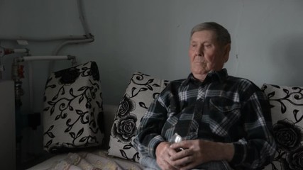 Old grandfather looking thoughtfully aside sits on the couch in the interior of a village apartment with glasses in his hands and watching TV - Powered by Adobe